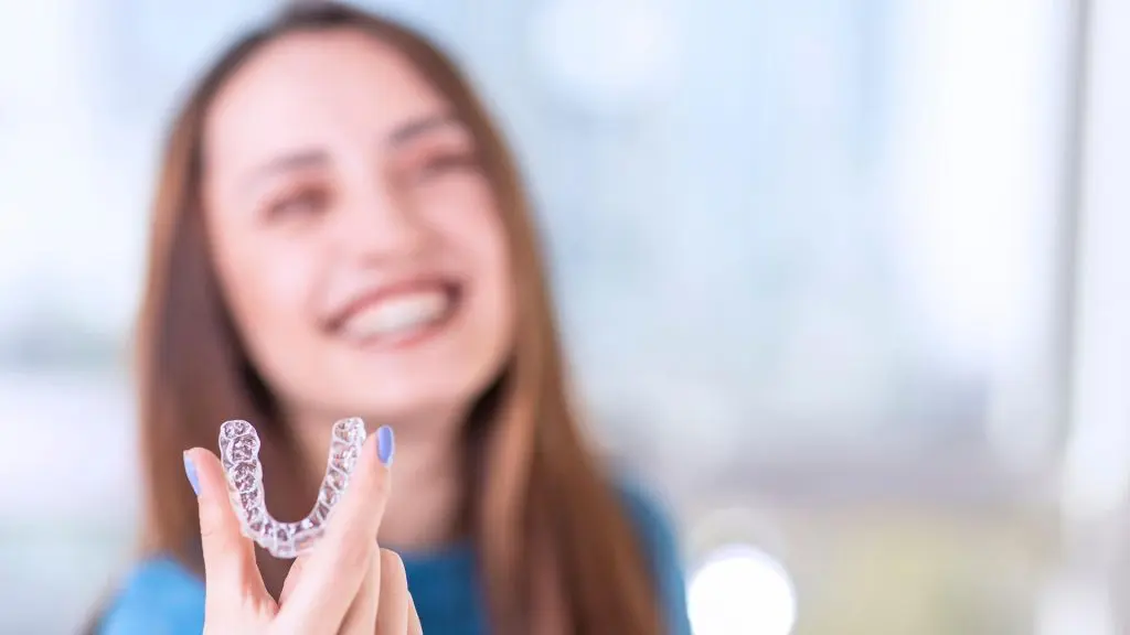 A blurred-out woman is holding a clear aligner up so you can see it.